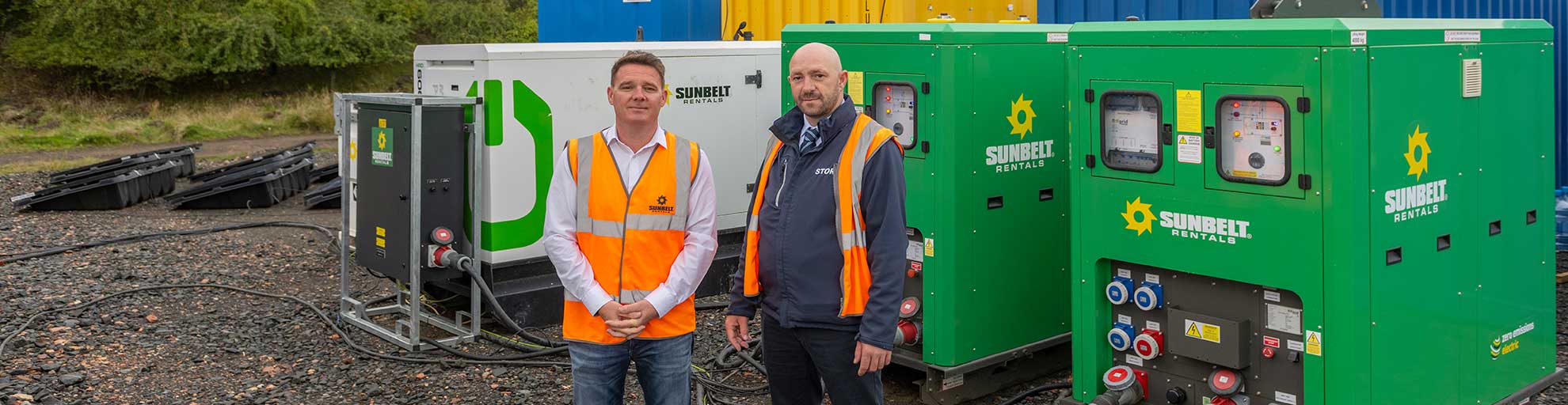 Two Project Managers Stood In front Of Battery Storage Units And A Hybrid Generator